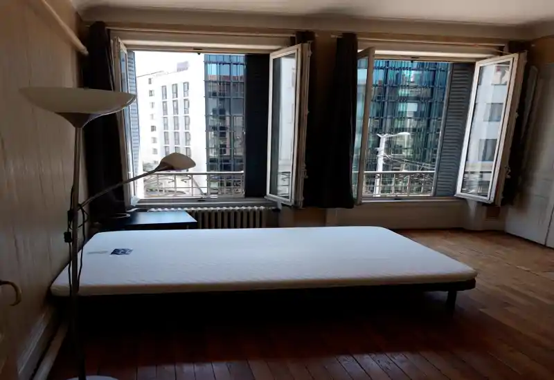 Bright bedroom with a large mattress centered near three open windows, hardwood floors and a standing lamp; ideal for showcasing natural light and city views.