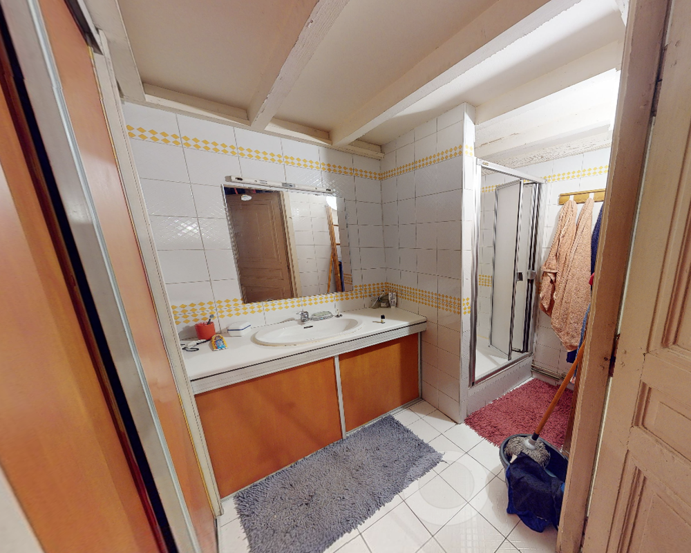 Salle de bain carrelée lumineuse avec un lavabo sur meuble, grand miroir et cabine de douche ; tapis et articles de toilette visibles, vue nette des installations.