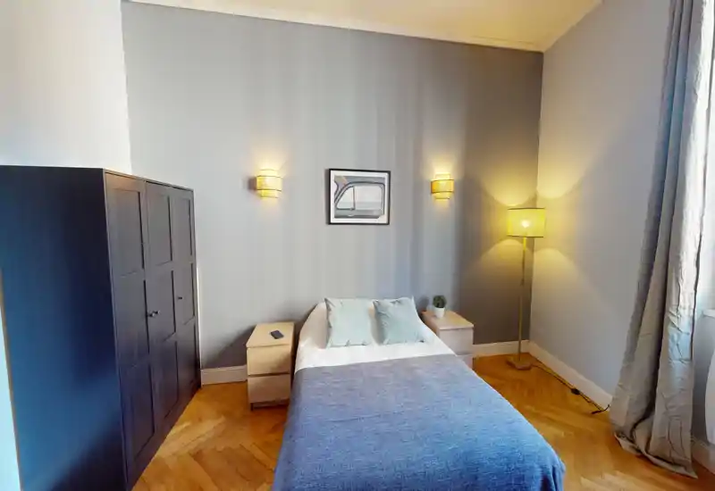 Camera da letto luminosa e ben arredata con letto matrimoniale centrato su una parete grigio chiaro, due comodini, applique e una lampada da terra vicino alla finestra.
