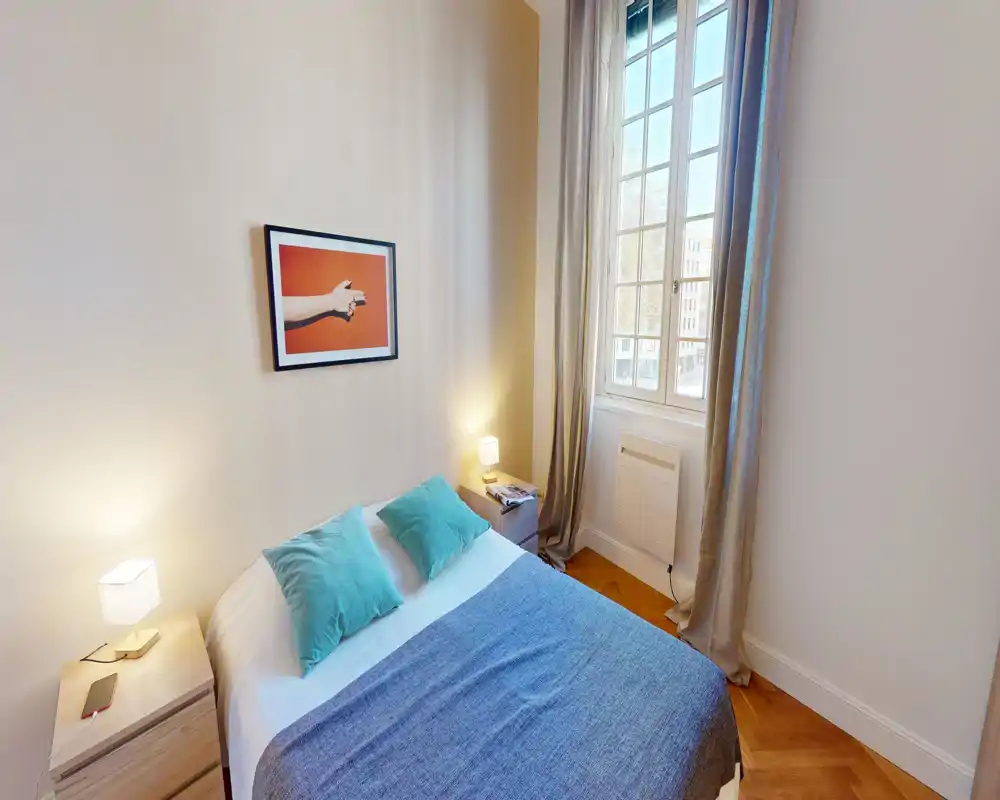 Camera da letto luminosa e curata con un letto matrimoniale con biancheria blu, due comodini con lampade e una grande finestra che illumina la stanza.