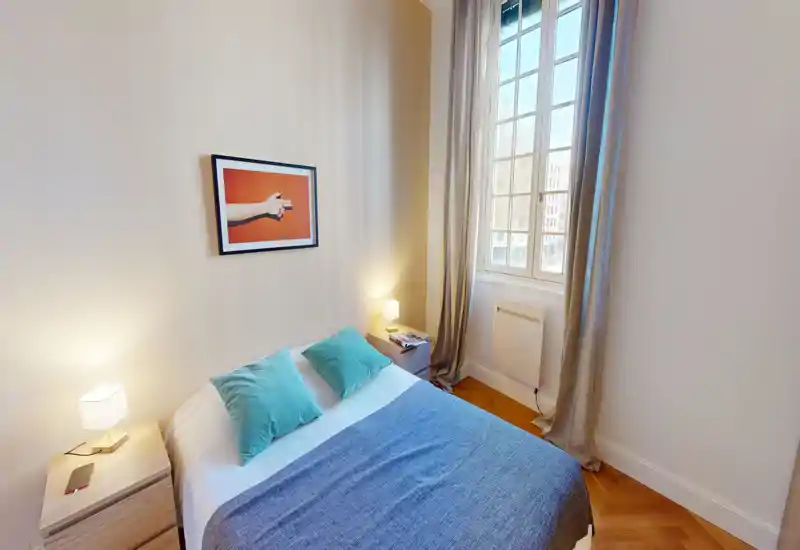Camera da letto luminosa e curata con un letto matrimoniale con biancheria blu, due comodini con lampade e una grande finestra che illumina la stanza.
