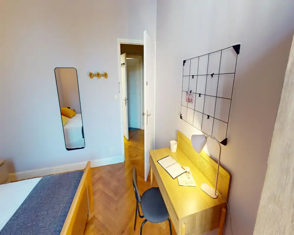 Chambre lumineuse et chaleureuse avec lit en bois et bureau soigné près d'une grille murale. Parquet chaleureux et linge doux pour une ambiance accueillante.