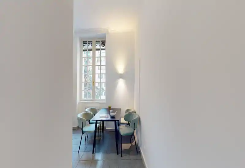 Bright, narrow dining area with a modern table and four upholstered chairs placed near a tall window; soft wall lighting creates a cozy minimalist atmosphere.