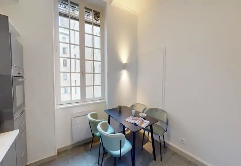 Small, stylish dining corner with a compact table and four velvet-upholstered chairs, lit by a tall window providing natural light.