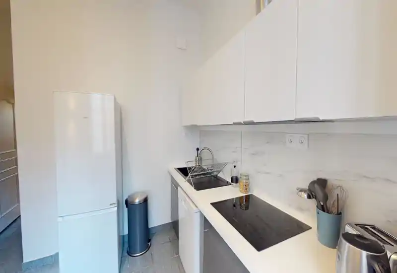 Cucina moderna compatta con pensili bianchi, piano ad induzione, frigorifero e piccoli elettrodomestici. Schienale effetto marmo e disposizione pratica ideale per affitti brevi.