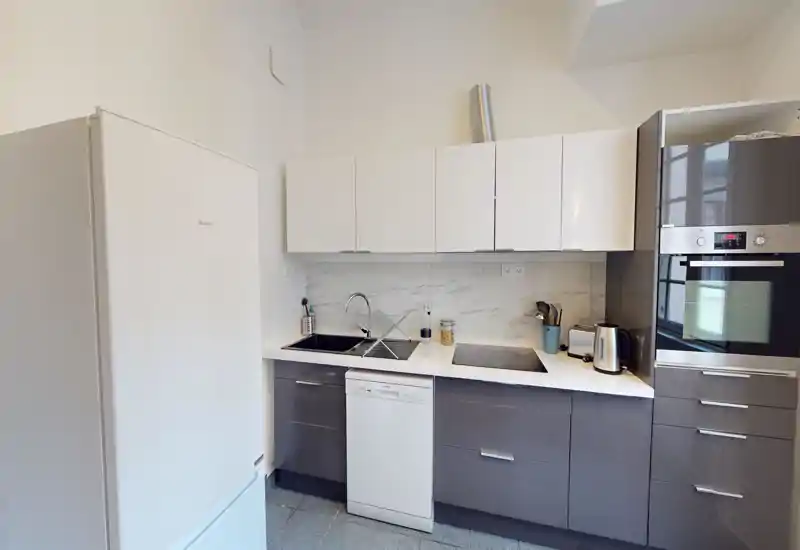Cucina moderna e compatta con forno integrato, frigorifero, lavastoviglie e doppio lavello su uno sfondo effetto marmo pulito.