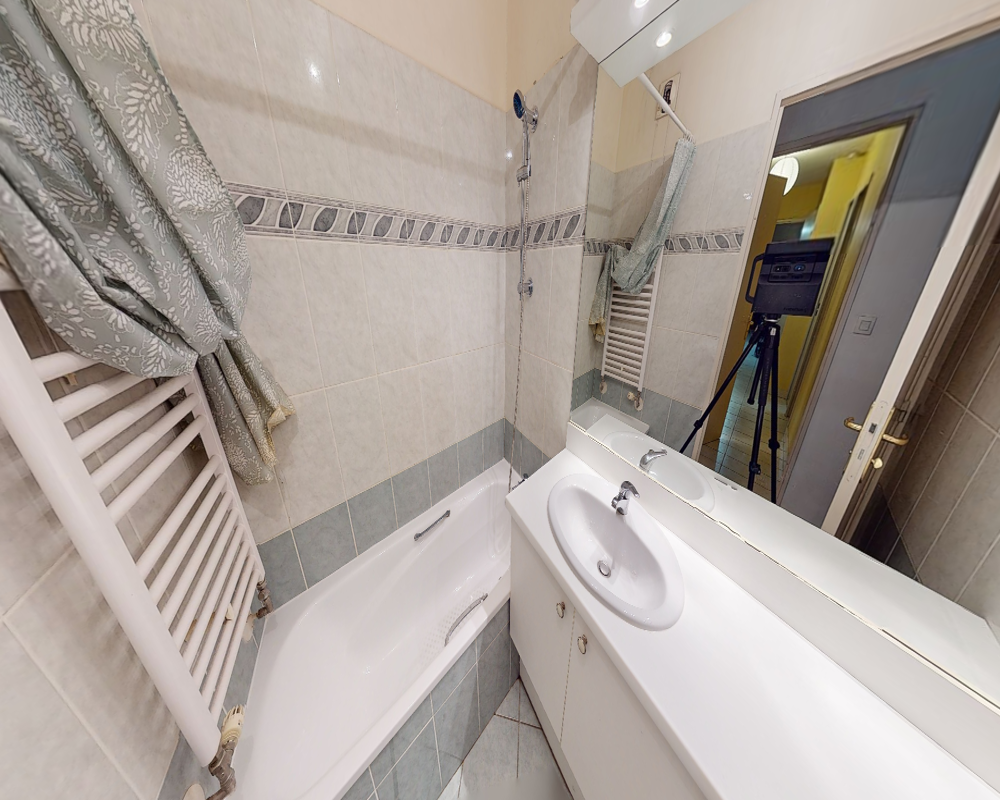 Salle de bains compacte et bien éclairée avec baignoire, pommeau de douche mural, lavabo avec meuble et radiateur serviette ; grand miroir reflétant le couloir d'entrée.