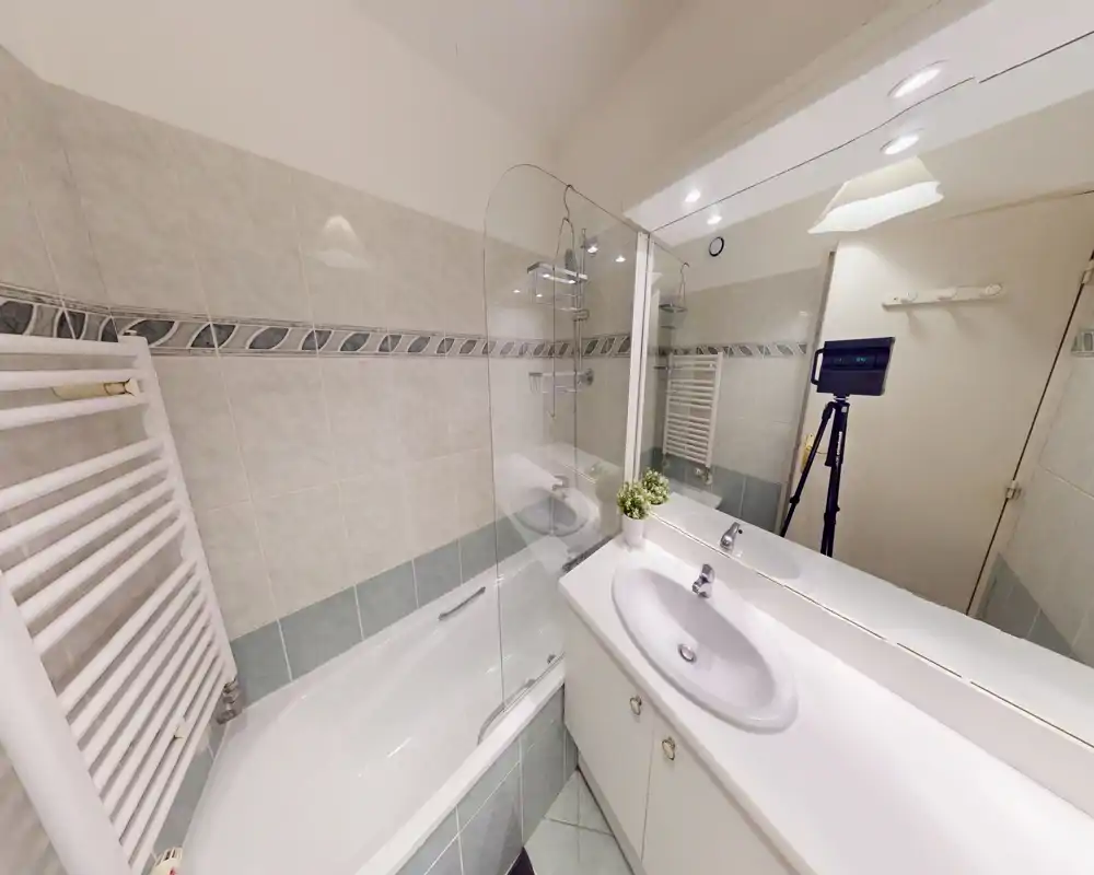 Salle de bains propre et lumineuse avec baignoire et douche, pare-douche en verre, lavabo simple avec grand miroir et meuble sous vasque ; radiateur sèche-serviettes visible.