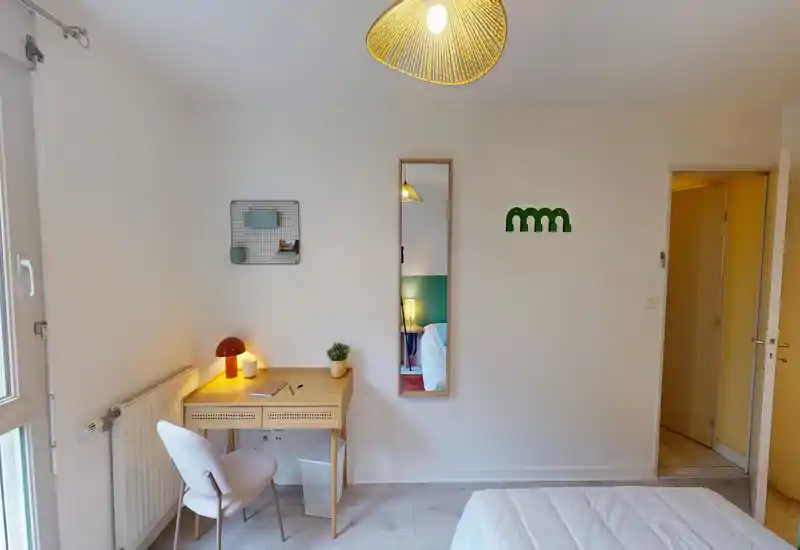 Piccola camera luminosa con bordo del letto visibile, una scrivania compatta in legno e sedia vicino alla finestra, lampada calda e specchio a tutta altezza che crea profondità.