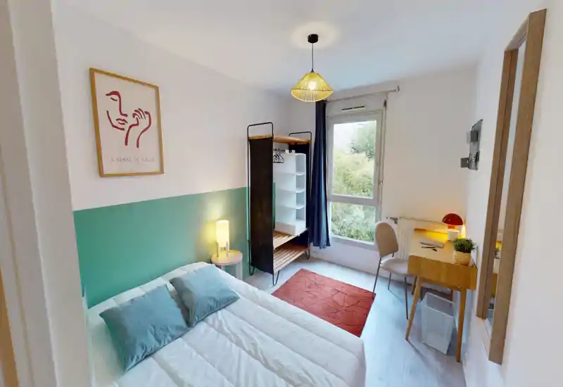 Camera da letto luminosa e accogliente con letto matrimoniale, lampada da comodino, guardaroba aperto e scrivania vicino alla finestra. Parete accentuata in verde acqua e tessili caldi creano un’atmosfera piacevole; ampia finestra con vista sul verde.