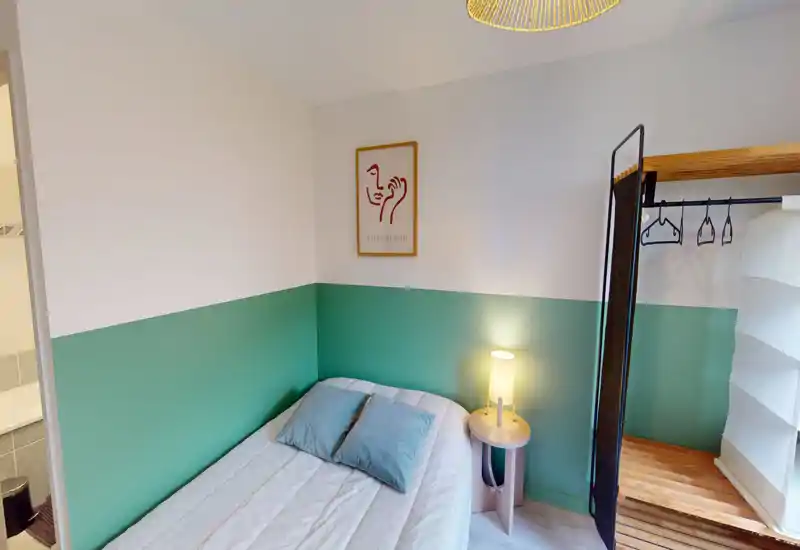 Camera da letto piccola e curata con letto matrimoniale, comodino con lampada, parete verde a mezza altezza e armadio aperto — luminosa e accogliente per soggiorni brevi.