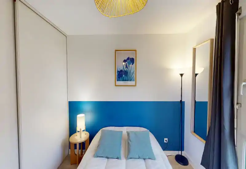 Cozy, well-lit bedroom with a centrally placed double bed dressed in white linens and light blue cushions, a bedside lamp, floor lamp and a full-length mirror that enhance the space.