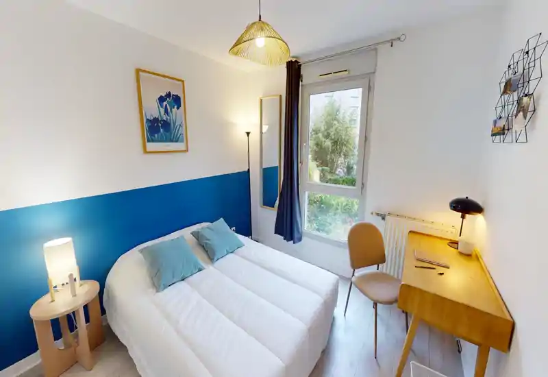 Bright, well-styled double bedroom with a neatly made bed, bedside lamp and a small wooden desk by the window overlooking greenery.