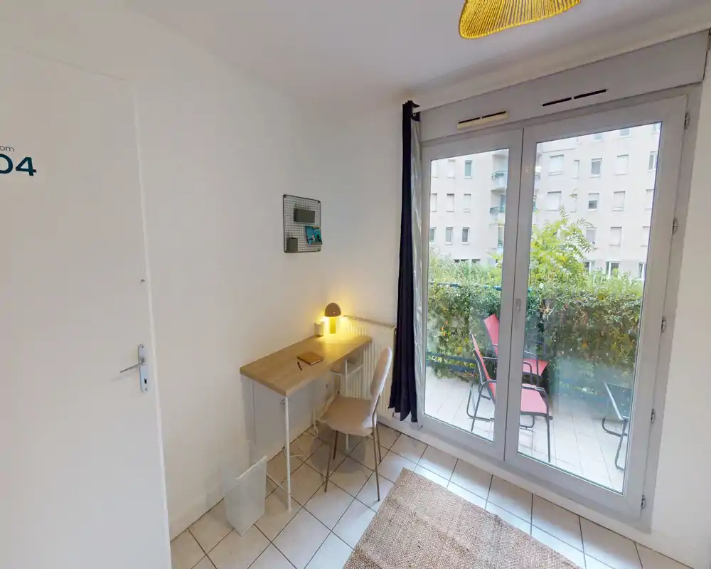 Compact bedroom corner with a small desk and chair next to large glass doors opening to a balcony. Bright, simply furnished space with a rug and lamp creating a cozy study nook.