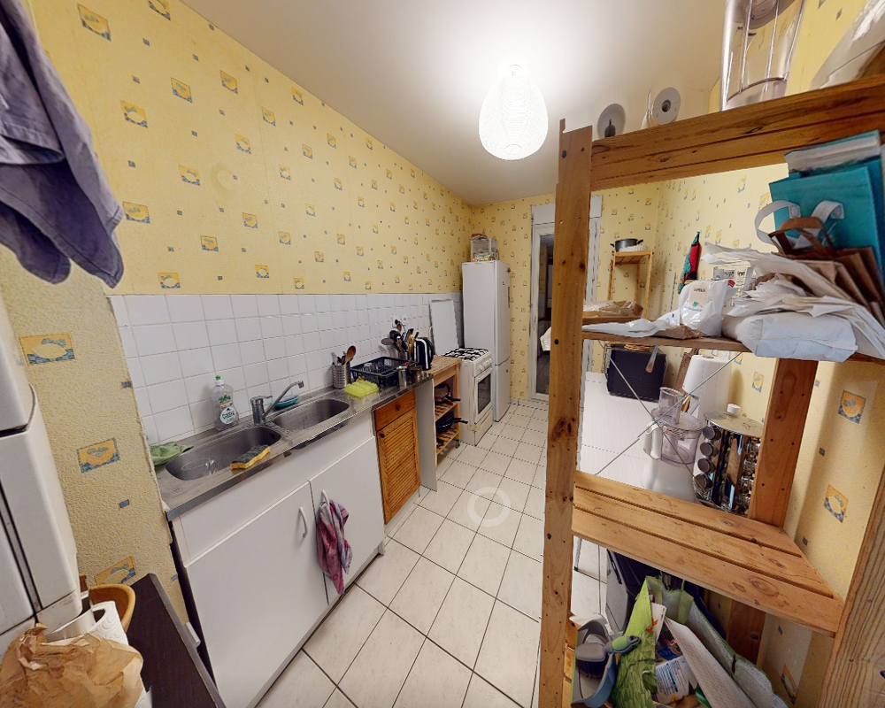 Cuisine en enfilade étroite avec évier, cuisinière et réfrigérateur indépendant ; étagères en bois pour la vaisselle et les provisions. Le papier peint jaune et le carrelage au sol apportent une atmosphère chaleureuse et fonctionnelle.