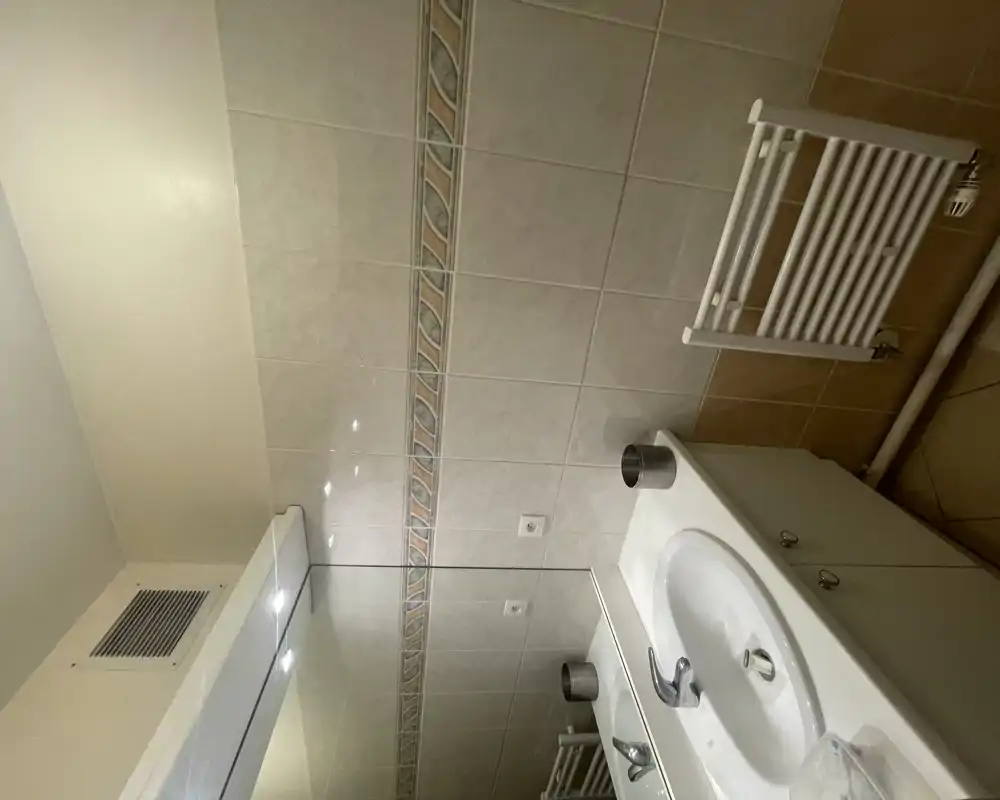 Salle de bain compacte et propre avec lavabo blanc, meuble sous-vasque, miroir et sèche-serviettes chauffant ; murs carrelés neutres avec une frise décorative.