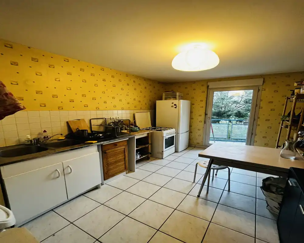 Cuisine fonctionnelle avec sol carrelé, évier sur plan de travail, cuisinière et réfrigérateur indépendants, table à manger et lumière naturelle provenant d'une porte vitrée donnant sur l'extérieur.