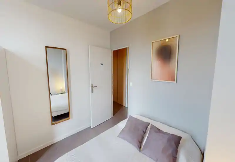 Compact, softly styled bedroom with a double bed dressed in neutral linens, two pillows and a full-length mirror beside the door; calm tones and a pendant light create a welcoming atmosphere.