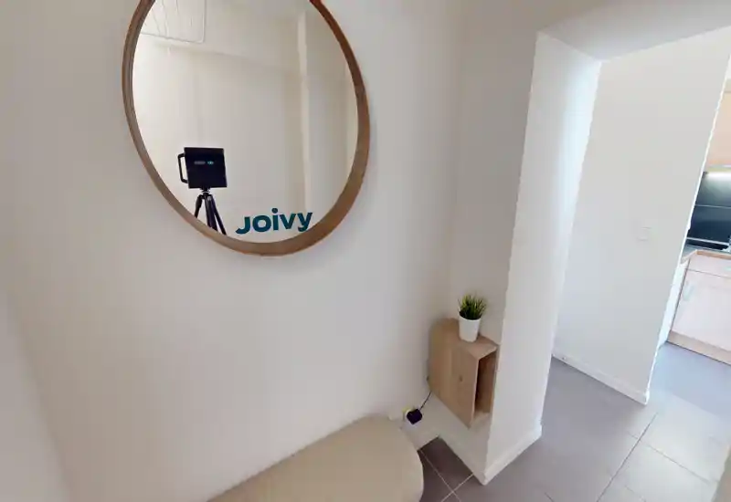 Entrée lumineuse et soignée avec un miroir rond, un petit banc et une étagère murale avec une plante ; sol carrelé menant à la cuisine.