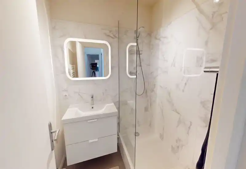Salle de bains moderne et compacte avec douche à l'italienne, miroir éclairé, meuble vasque et carrelage effet marbre — lumineuse et bien entretenue.