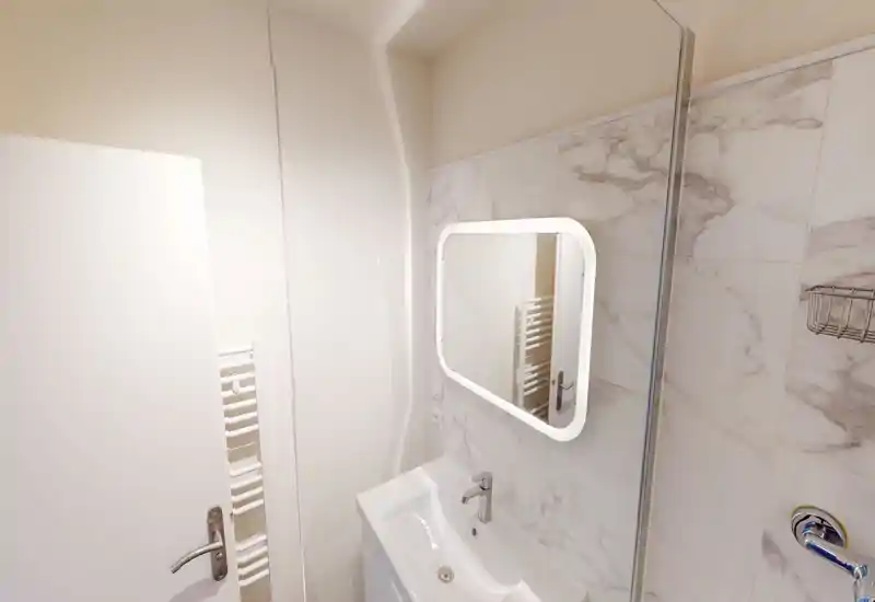 Salle de bain moderne et compacte avec lavabo mural, miroir éclairé et carreaux imitation marbre — lumineuse et propre.