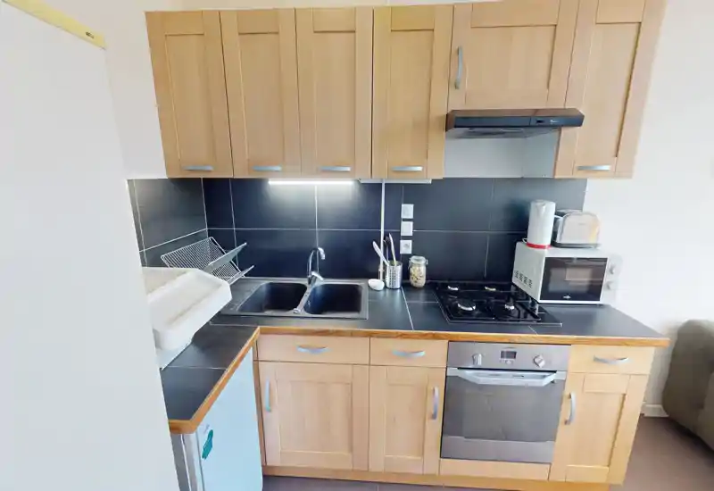Cuisine compacte bien équipée avec placards en bois, évier double, plaque à gaz et four encastré — agencement lumineux et soigné, idéal pour un petit appartement.
