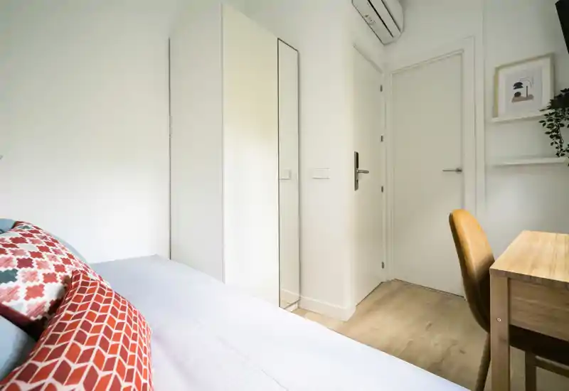 Camera da letto luminosa e compatta con letto ben rifatto, cuscini decorativi e una piccola scrivania. Armadio bianco con specchio a tutta altezza e pavimento in legno chiaro creano un ambiente fresco e arioso, perfetto per una persona o soggiorni brevi.