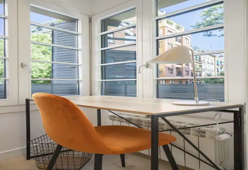 Angolo luminoso e moderno con scrivania e sedia arancione davanti a grandi finestre — perfetto come studio domestico o angolo lettura con molta luce naturale.