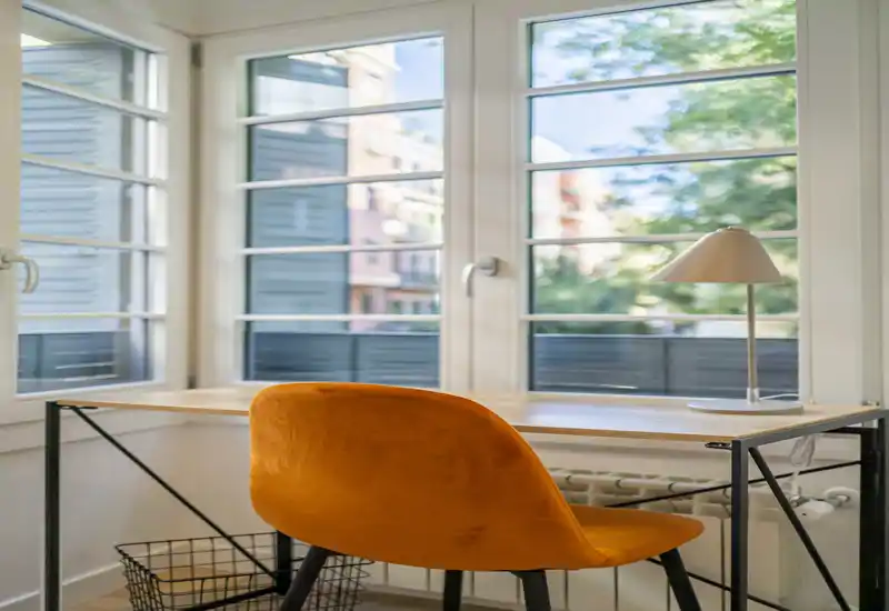 Angolo di lavoro luminoso con scrivania in legno moderna, sedia imbottita arancione e lampada da tavolo davanti a grandi finestre — ideale per smart working.