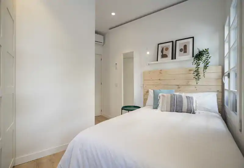 Bright, modern bedroom with a wooden headboard, crisp white bedding and decorative cushions; a small side table and wall art complete the cozy, minimalist look.