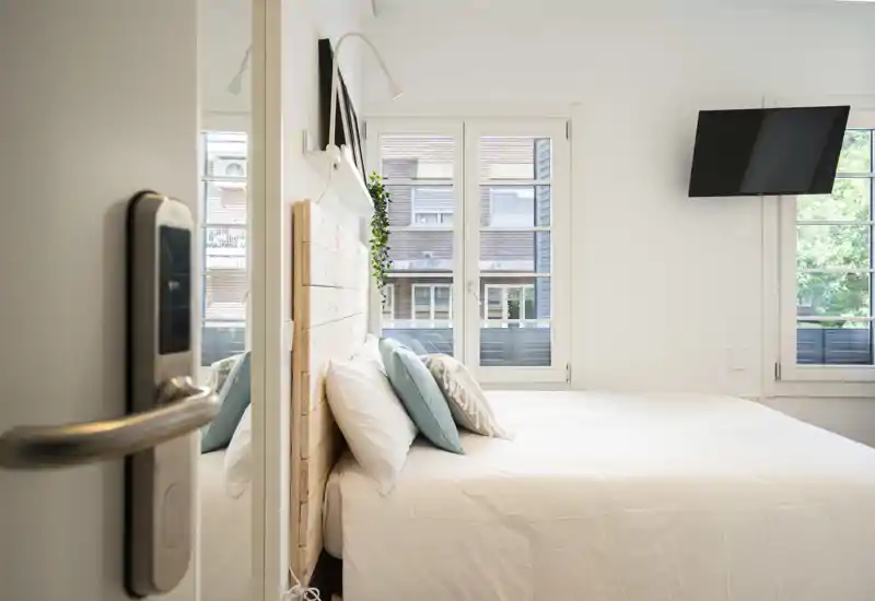 Bright, modern bedroom with a wooden headboard, crisp white bedding and soft accent pillows. Large windows provide abundant natural light and a wall-mounted TV keeps the space uncluttered.