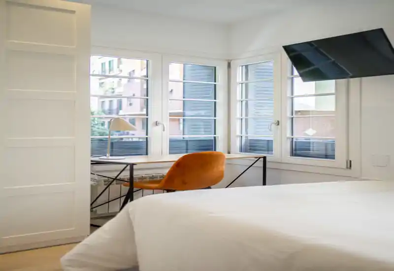 Camera da letto luminosa e moderna con ampio letto in primo piano, una scrivania davanti a tre finestre e una poltroncina arancione che crea un angolo lavoro accogliente.