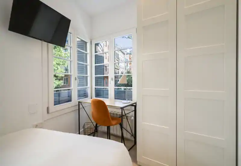 Camera compatta e luminosa con letto visibile, una piccola scrivania vicino alle grandi finestre e un armadio bianco su misura — ideale per una persona o soggiorni brevi.