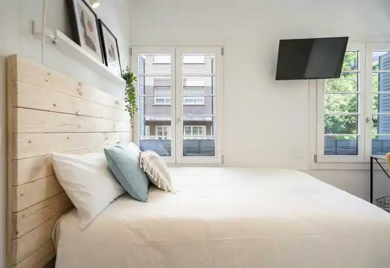 Bright, modern bedroom with a wooden headboard, crisp white bedding and decorative cushions. Large windows fill the room with natural light and a wall-mounted TV is visible.