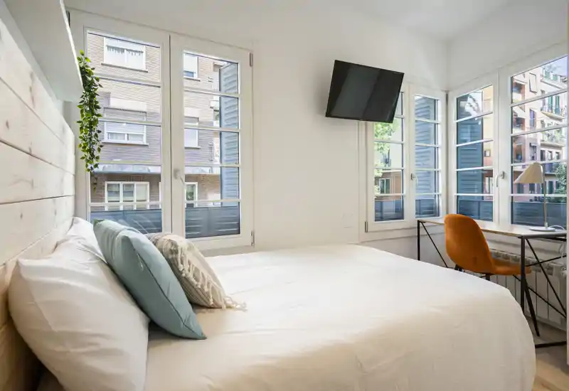 Bright, modern bedroom with a large bed, wood headboard and a desk by multiple windows letting in abundant natural light.