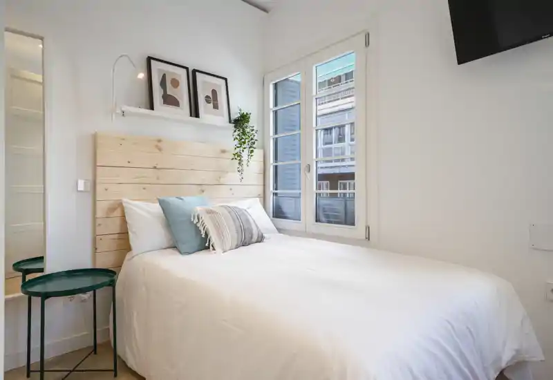 Bright, neatly styled bedroom with a white bed, wooden headboard, decorative cushions and natural light from the window.