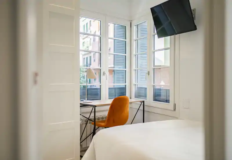 Angolo di camera luminoso e moderno con letto bianco, ampie finestre e una scrivania accogliente con sedia arancione — perfetto per lavorare o rilassarsi con molta luce naturale.