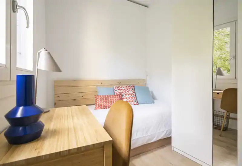 Chambre lumineuse et compacte avec tête de lit en bois, coussins décoratifs et un petit espace de travail près de la fenêtre — bien éclairée et soigneusement mise en scène.