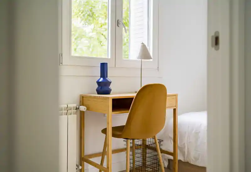 Angolo camera luminoso e minimale con scrivania e sedia in legno accanto a una finestra; si intravede il letto con biancheria bianca e luce naturale calda.