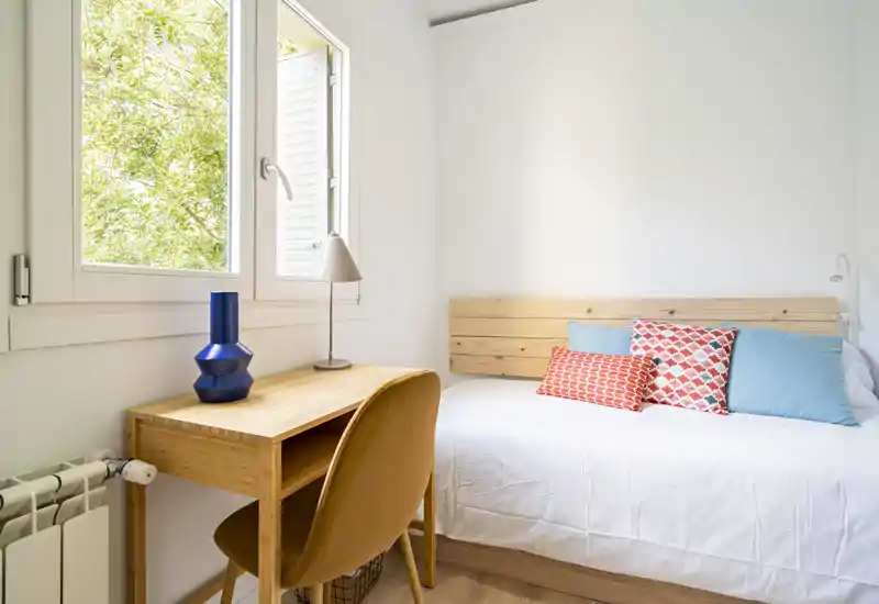 Chambre lumineuse et minimaliste avec tête de lit en bois et coussins à motifs. Un petit bureau près de la fenêtre apporte de la lumière naturelle, idéal pour travailler ou se détendre.