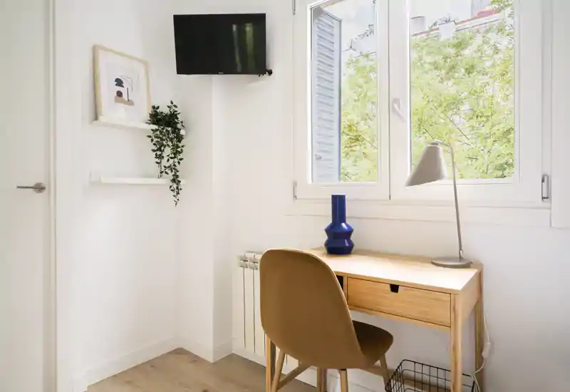 Angolo studio luminoso e minimal in un soggiorno: scrivania in legno vicino alla finestra, sedia confortevole, vaso decorativo e pianta a parete, perfetto per il lavoro da casa.