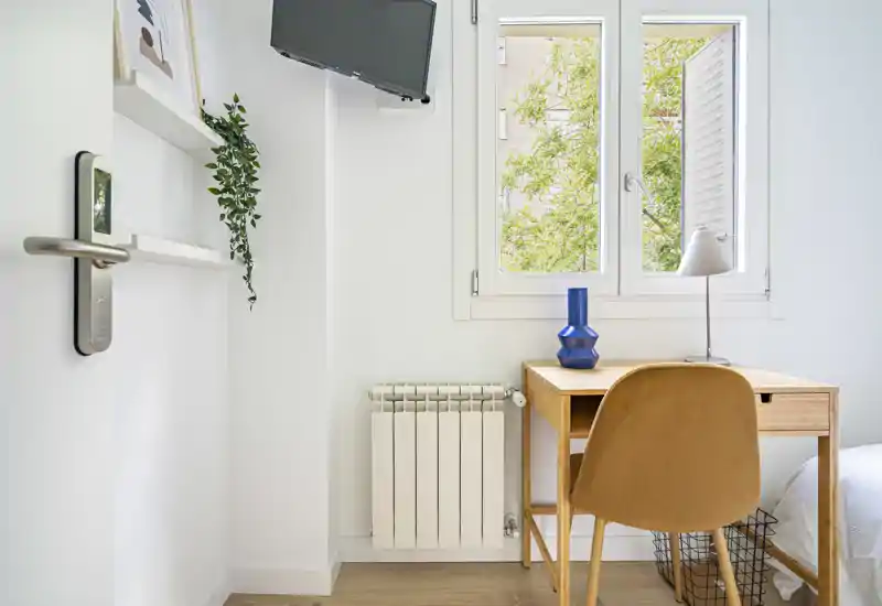 Angolo di camera luminoso e compatto con una scrivania in legno semplice vicino alla finestra, una parte del materasso visibile sul lato destro e una TV a parete. Luce naturale e arredi minimalisti per un’atmosfera pulita e ariosa, adatta a un affitto breve o per studenti.