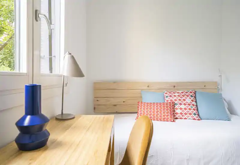 Chambre lumineuse et minimaliste avec tête de lit en bois, coussins décoratifs et petit bureau près de la fenêtre — idéale pour un séjour cosy et bien éclairé.