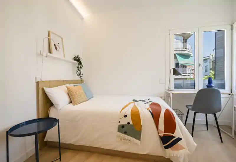 Chambre simple lumineuse et chaleureuse avec tête de lit en bois, jeté coloré et petit bureau près de la fenêtre — idéale pour un séjour confortable ou un étudiant.