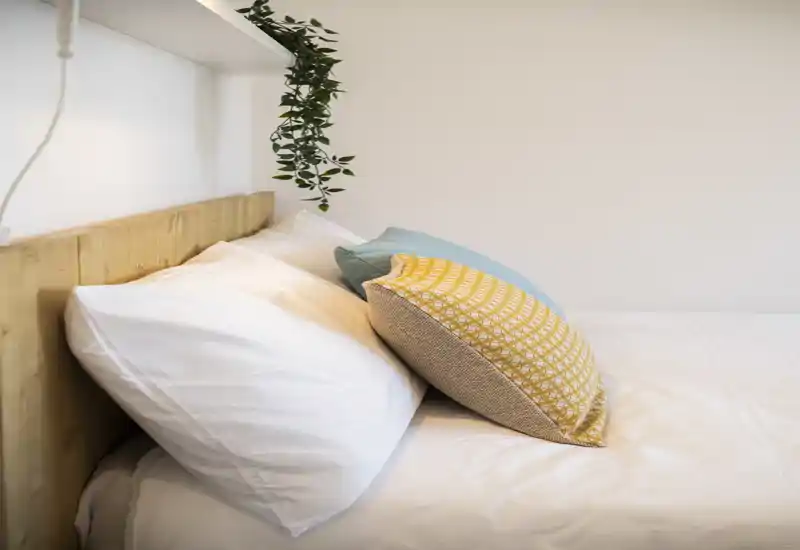Coin chambre lumineux et minimaliste avec linge de lit blanc immaculé, coussins décoratifs superposés et tête de lit en bois clair — chaleureux et bien mis en scène pour une annonce.