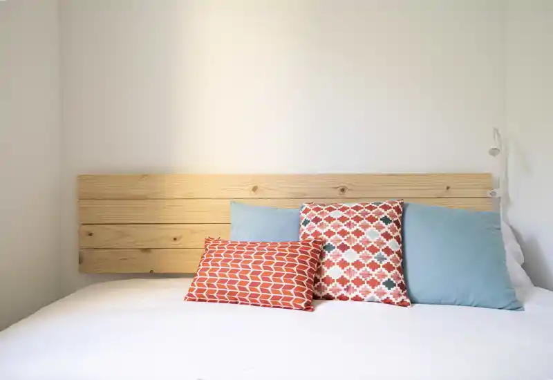 Bright, minimalist bedroom with a wooden slat headboard and decorative cushions on a white bedspread, offering a fresh and welcoming sleeping area.