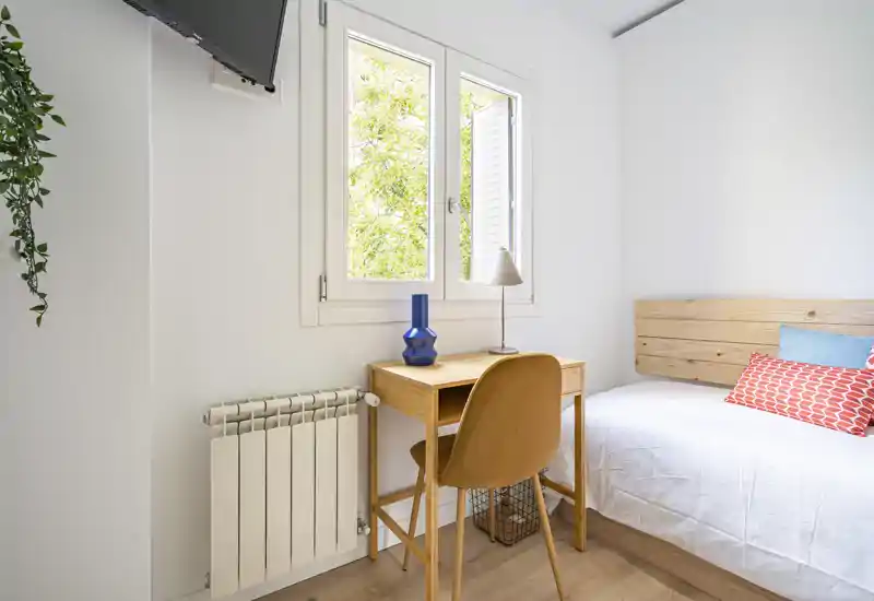 Bright, compact bedroom with a single bed, wooden headboard and a small desk by the window; fresh white walls and colorful cushions create a neat, welcoming feel.