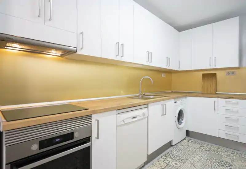 Cocina moderna y luminosa con armarios blancos, encimeras de madera, electrodomésticos integrados y suelos con baldosas decorativas — limpia y lista para usar.