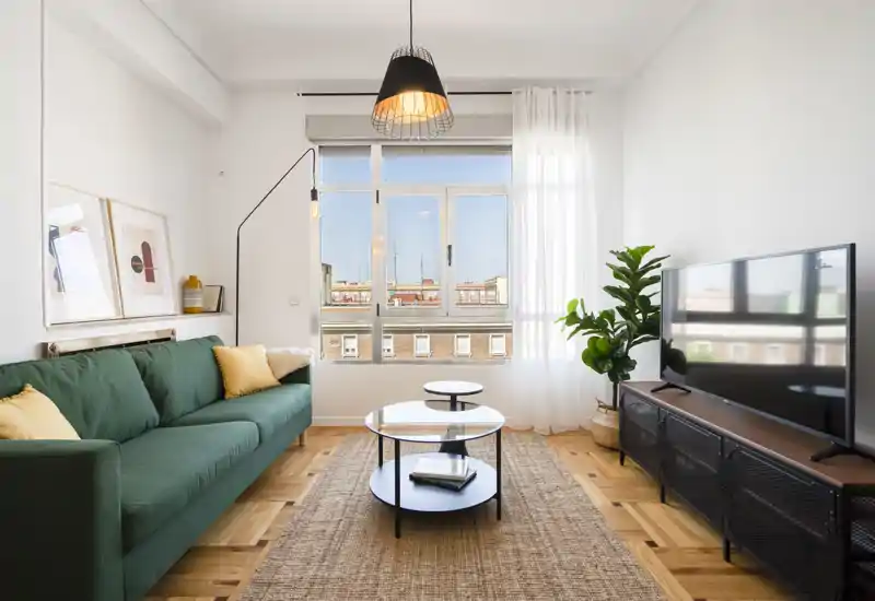 Salón luminoso y moderno con sofá verde, gran ventana que aporta luz natural y mueble TV — alfombra y plantas crean un ambiente acogedor.