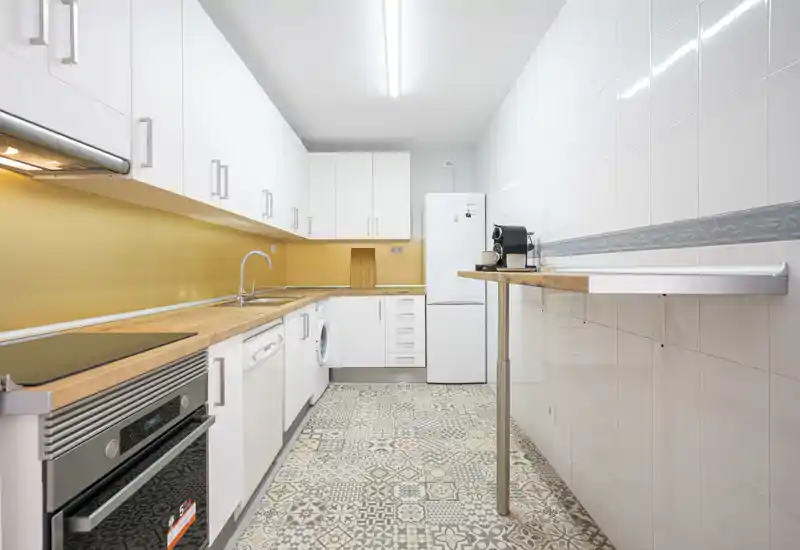 Cocina tipo galería, moderna y luminosa con armarios blancos, encimeras de madera y suelo de baldosas decoradas; equipada con horno, fregadero, nevera y lavadora — ordenada y lista para usar.